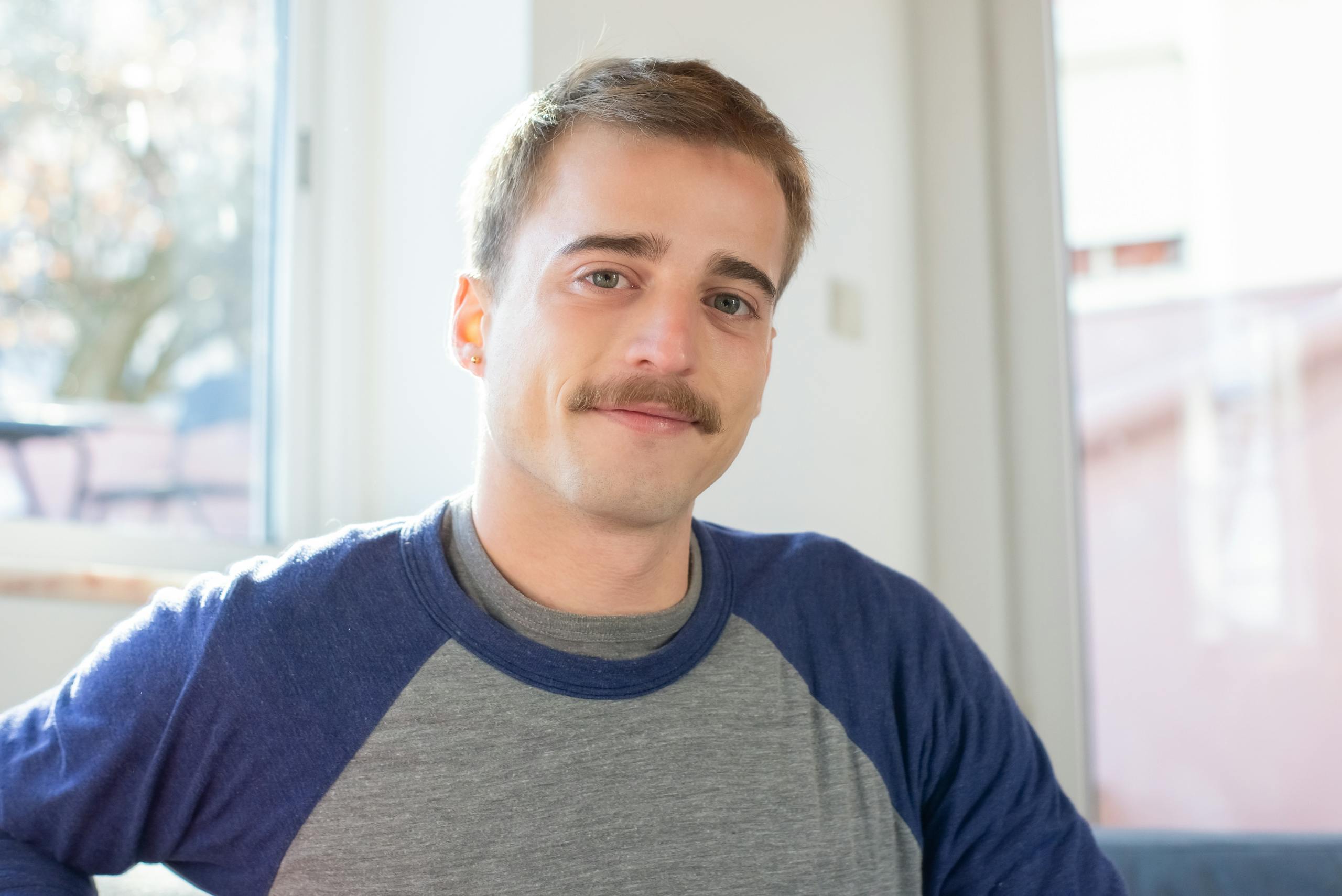 A relaxed portrait of a smiling man with a mustache indoors, wearing a casual blue and gray shirt.