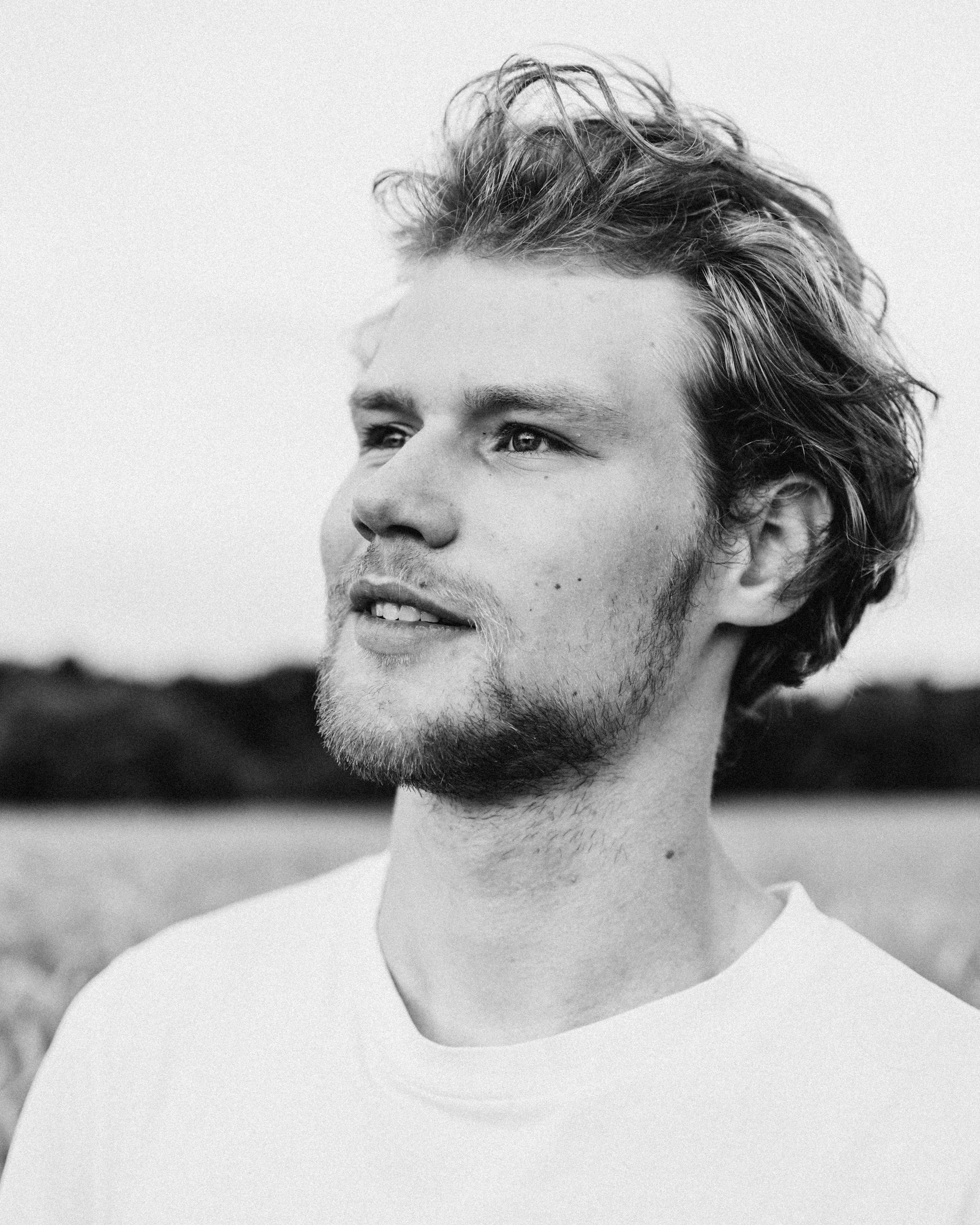 Captivating black and white portrait of a young bearded man looking away, wearing a white t-shirt outdoors.
